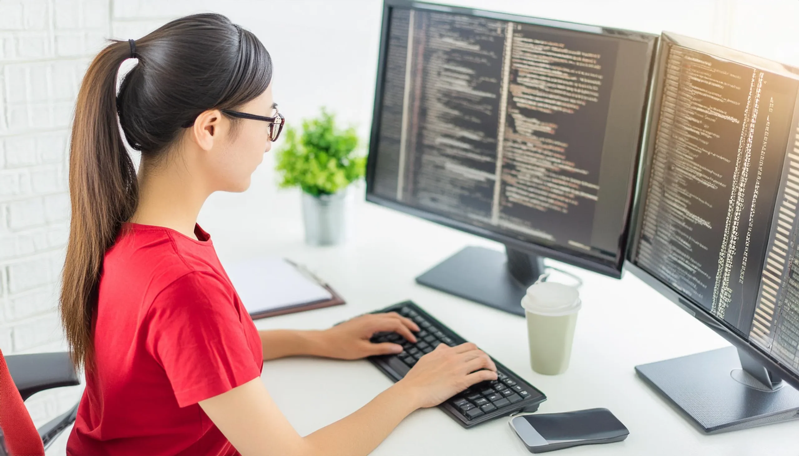 プログラミングをしている日本人の女性。赤いTシャツ。背景はプログラミングコード。横からの風景